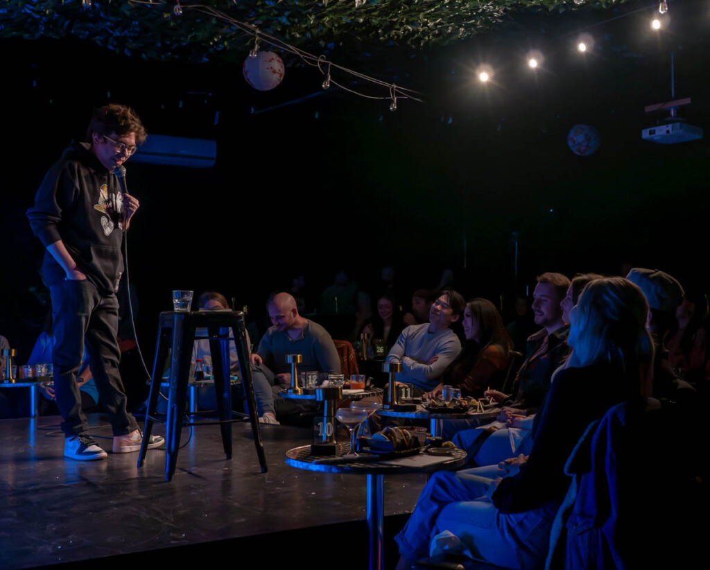 A live comedy show at the Denver Comedy Lounge. A comedian tells a joke as the participants snack on pork bao buns during the show between laughs.