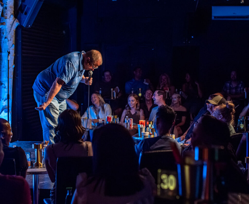 A stand-up comedian bowing in front of a laughing audience at the Denver Comedy Lounge club.