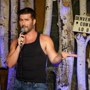 Comedian Corey O'Brien Live performing on stage at the Denver Comedy Lounge. Corey wearing a black tank top with his perfectly cut beard and dark hair performs on stage in front of the iconic Denver Comedy Lounge aspen tree backdrop. This image promotes his upcomeing comedy show at the Denver Comedy Lounge on February 5th 2026