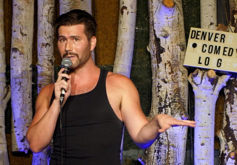 Comedian Corey O'Brien Live performing on stage at the Denver Comedy Lounge. Corey wearing a black tank top with his perfectly cut beard and dark hair performs on stage in front of the iconic Denver Comedy Lounge aspen tree backdrop. This image promotes his upcomeing comedy show at the Denver Comedy Lounge on February 5th 2026